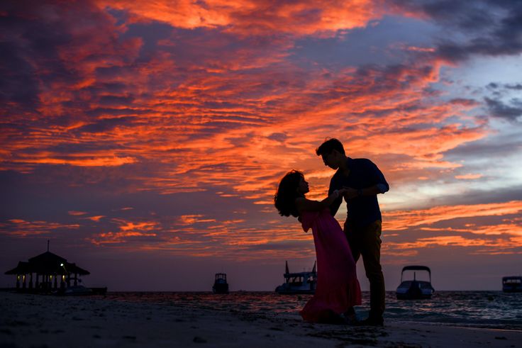 a man and woman standing on top of a sandy beach under a colorful sky at sunset