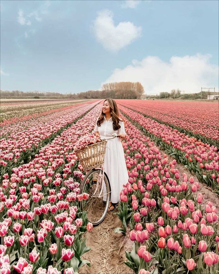 a woman in a white dress holding a basket standing next to a bike in a field full of pink tulips