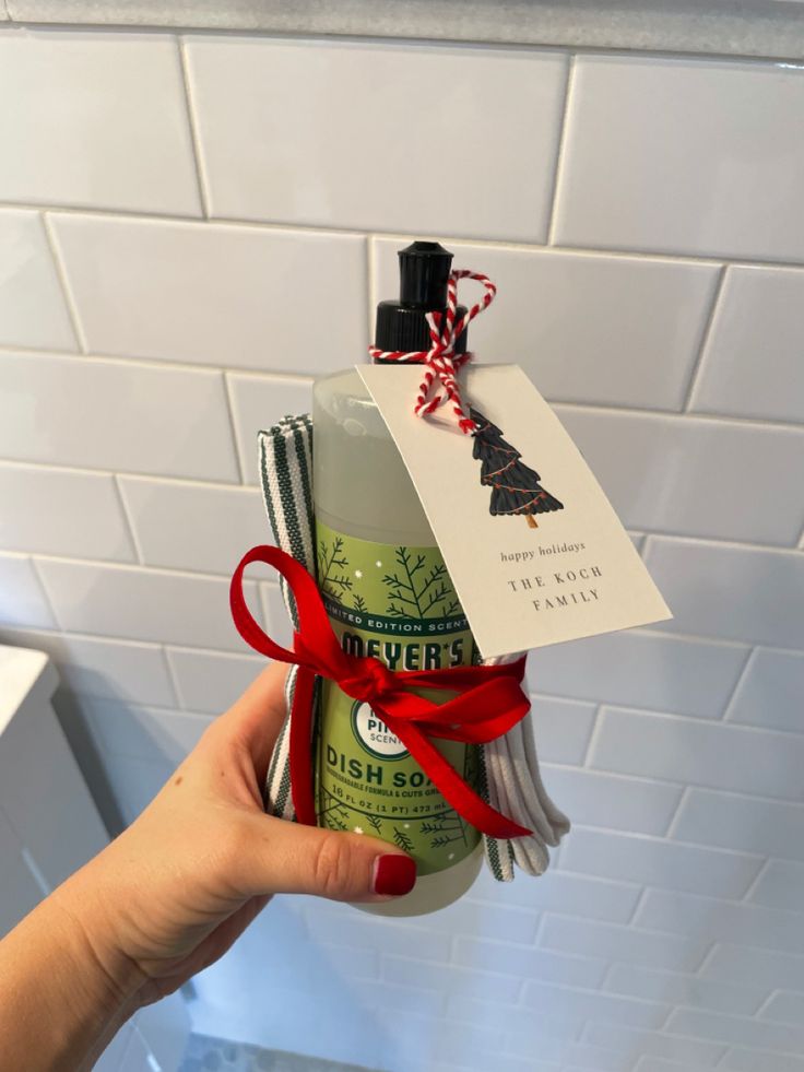 a hand holding a bottle of soap with a red ribbon tied around it in front of a white tiled wall