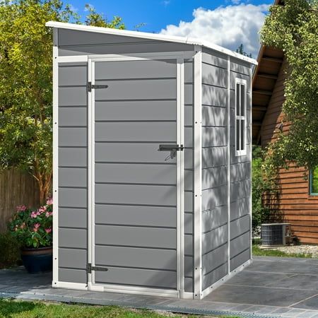 a small gray shed sitting on top of a yard next to a wooden fence and trees