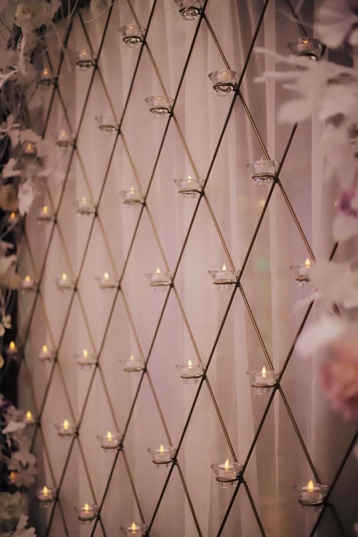 candles are lit on the side of a wall with flowers and curtains in the background