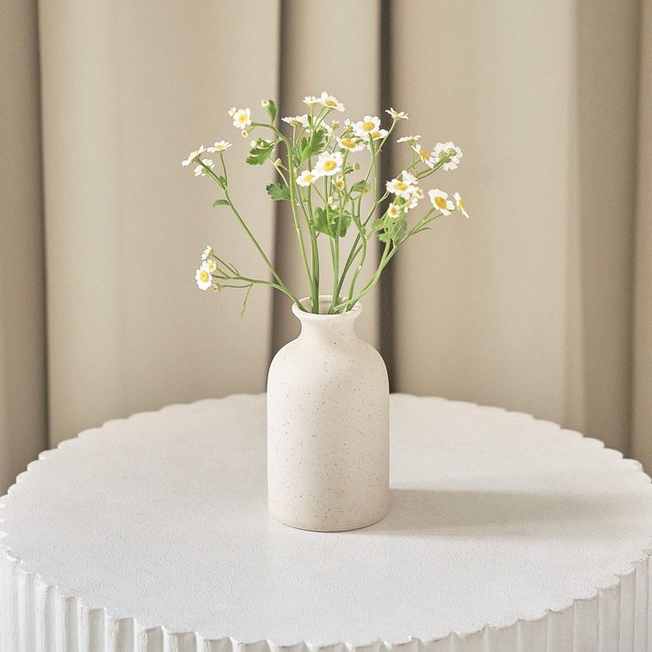 a white vase filled with flowers sitting on top of a round table next to a curtain