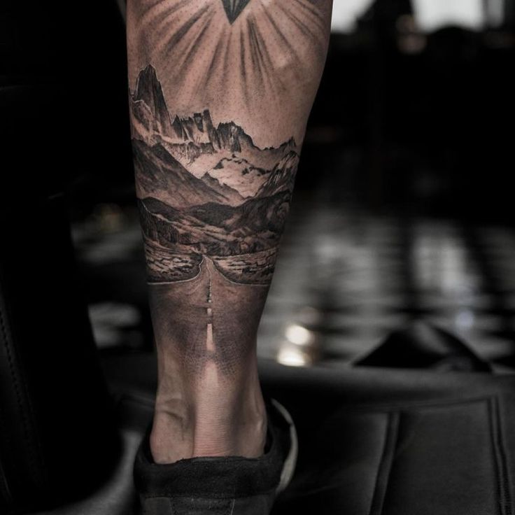 a man's leg with mountains and a cross tattoo on his left calf area