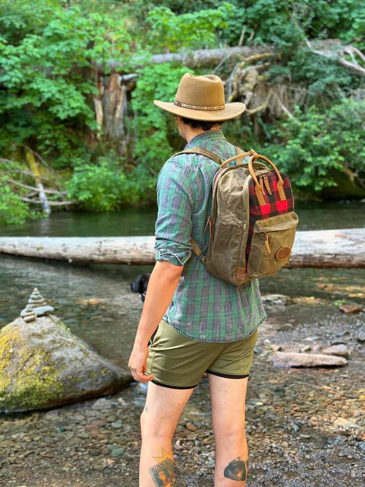 a person with a backpack and hat standing in front of a river, looking at the water