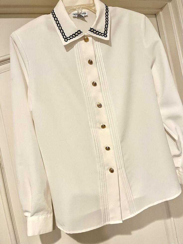 This is a beautiful classic 1990's soft-white wrinkle polyester perfectly tailored blouse with a row of 4 tucks on each side of the opening.  Beautiful metal gold buttons down the front and braided embroidery navy trip to the collar.  Travel in style with no-wrinkle long sleeves.  Chest measures 22 inches across at armpits and length 26 inches. Several marks to left of collar...see last photo.  This is new with tags and extra metal gold button. Mailed priority. White Semi-formal Blouse With Button Cuffs, White Blouse With Button Cuffs For Semi-formal Occasions, Elegant Tops With Peter Pan Collar And Buttons, Elegant Embroidered Shirt For Fall, Elegant Embroidered Fall Shirt, Formal Embroidered Button-up Shirt, Classic Embroidered Blouse For Work, Formal Blouse With Peter Pan Collar And Buttons, White Embroidered Formal Top