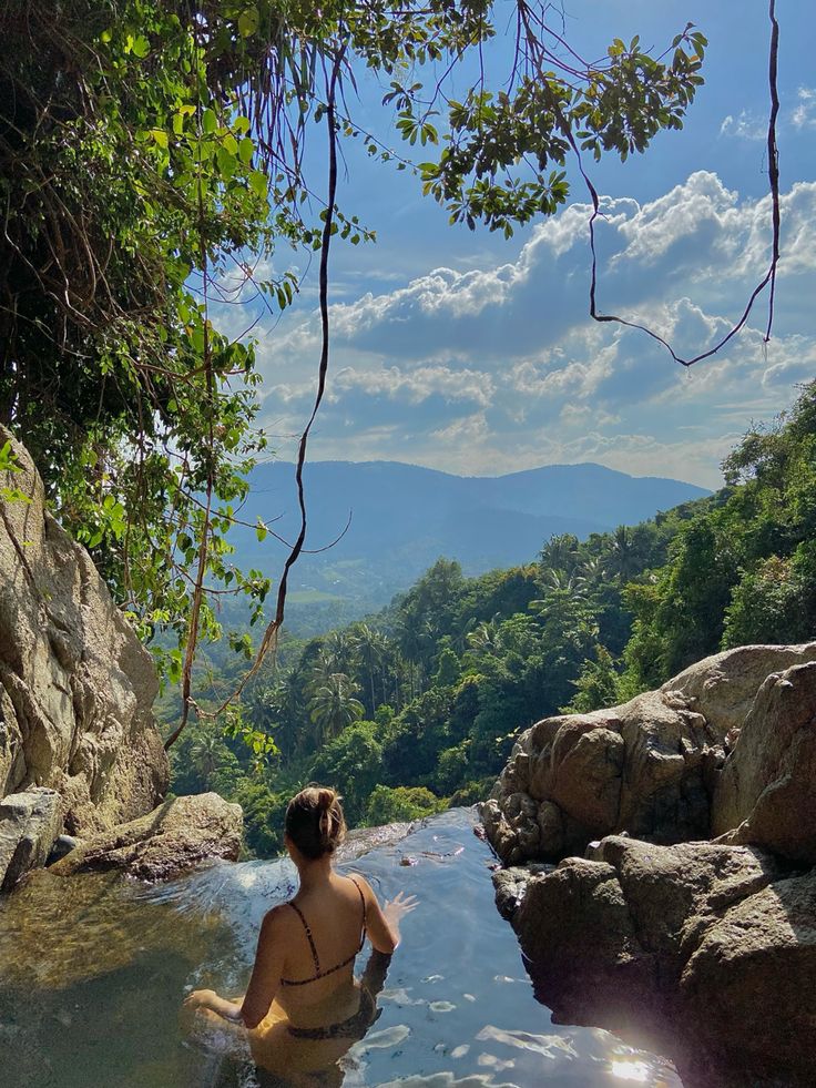 a woman sitting in the middle of a river with rocks and trees around her feet