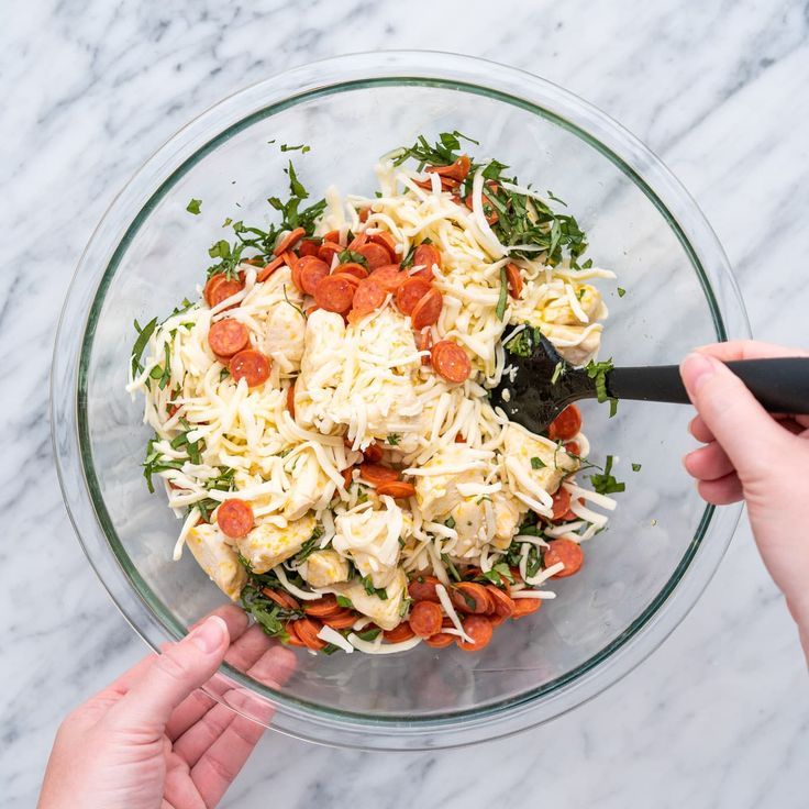 two hands holding a black spoon over a bowl of pasta and veggie salad