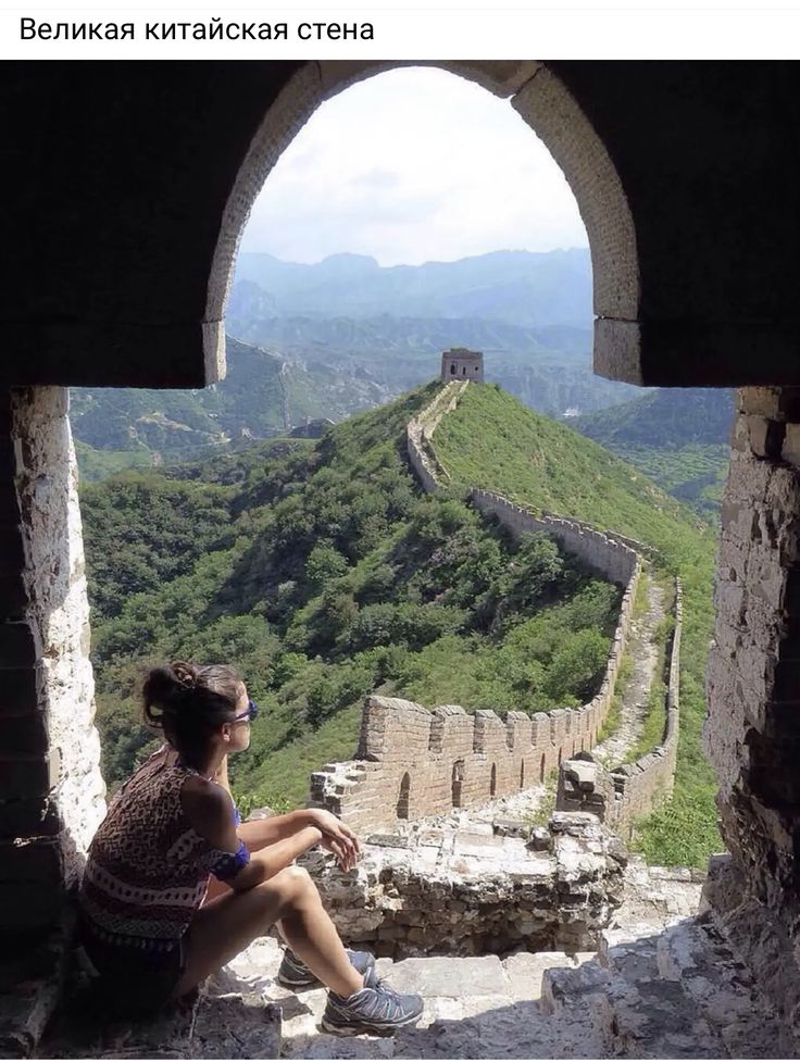 a woman sitting on top of a stone wall next to a green mountain covered in trees