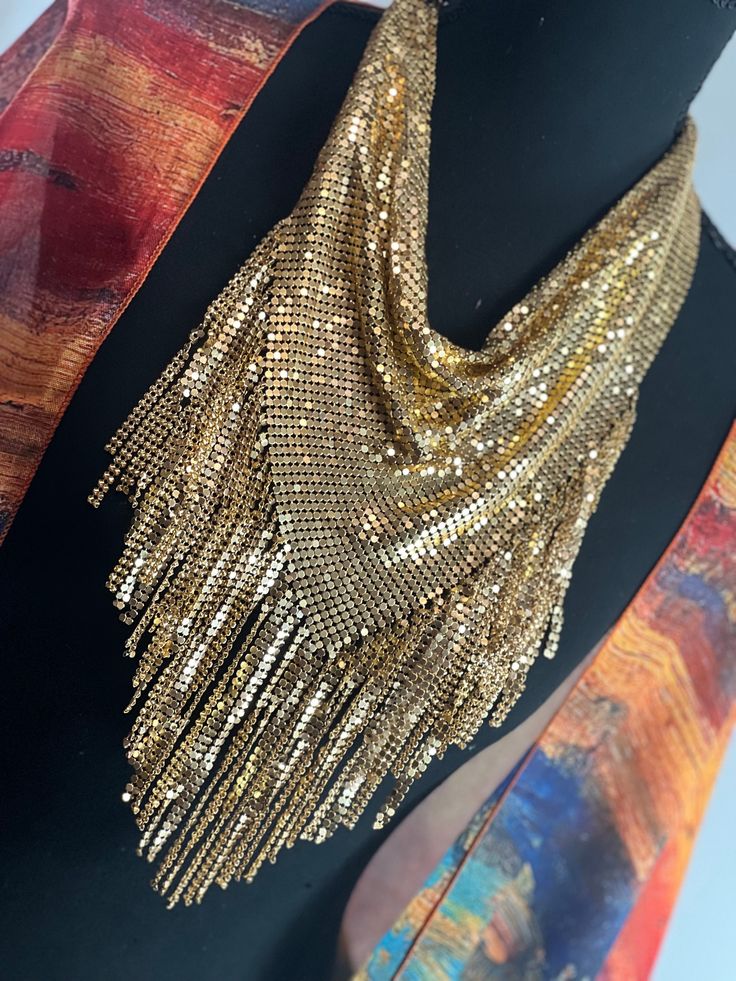 ### Gold Metal Mesh 1970s Style Bandana Necklace Scarf - Disco-Inspired Body Jewelry Embrace the glamour and sparkle of the 1970s with our Gold Metal Mesh Bandana Necklace Scarf. This dazzling piece combines the bold style of a bandana with the elegance of body jewelry, creating a statement accessory perfect for any disco-inspired look. ### Features: - **Design Triangular cowl design reminiscent of 1970s disco fashion, crafted to drape elegantly around the neck and shoulders. - **Materials Shimmering gold metal mesh that catches the light beautifully, adding a touch of vintage glamour. - **Versatile Wear Can be worn as a necklace, scarf, or body jewelry, making it a versatile addition to your accessory collection. - **Comfort Lightweight and flexible mesh material ensures a comfortable fit Gold-tone Chain Jewelry For Party, Glamorous Festival Jewelry With Chain Detail, Bohemian Party Chain Necklace With Adjustable Chain, Glamorous Metal Jewelry For Festival, Vintage Choker Chain Necklace For Party, Vintage Jewelry With Adjustable Chain For Party, Vintage Party Jewelry With Adjustable Chain, Bohemian Party Necklace With Adjustable Chain, Bohemian Necklace With Adjustable Chain For Party