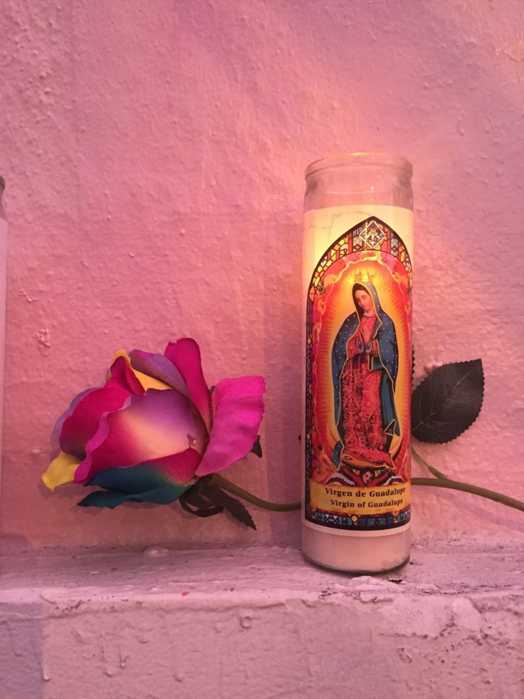 a lit candle with the image of mary on it next to a pink rose flower