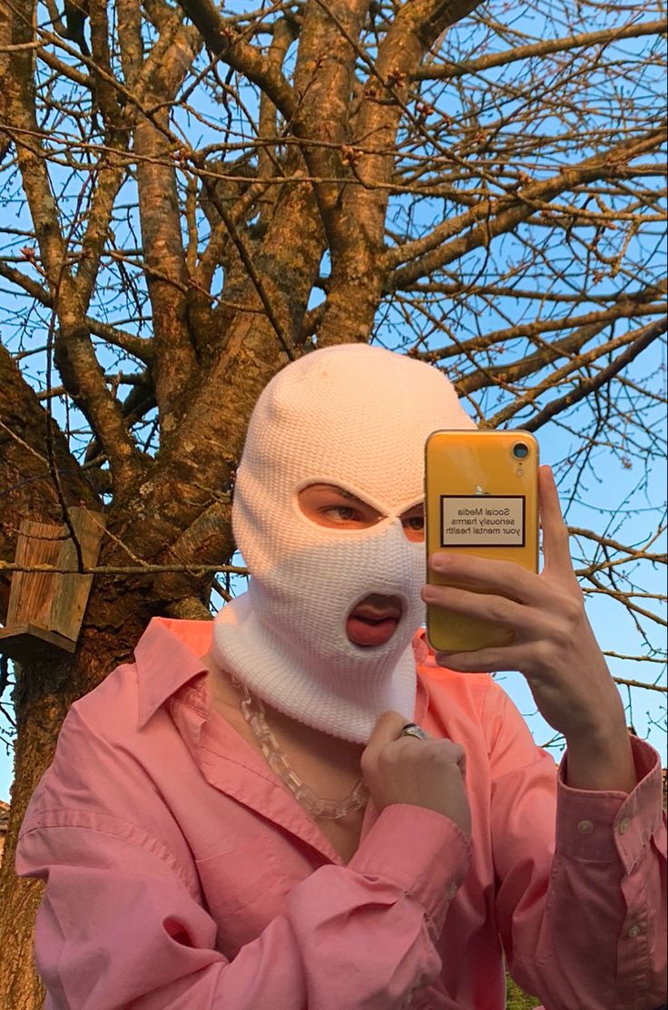 a woman wearing a white mask and holding up a cell phone to her face while standing in front of a tree