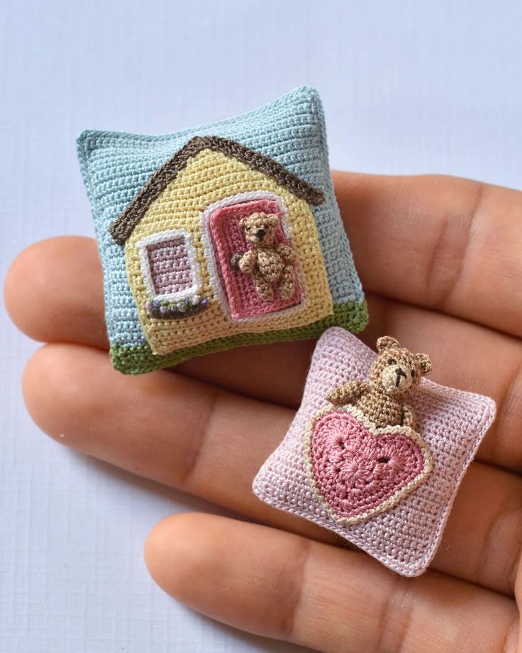 two small crocheted houses are sitting on someone's hand, one has a teddy bear and the other is a pillow