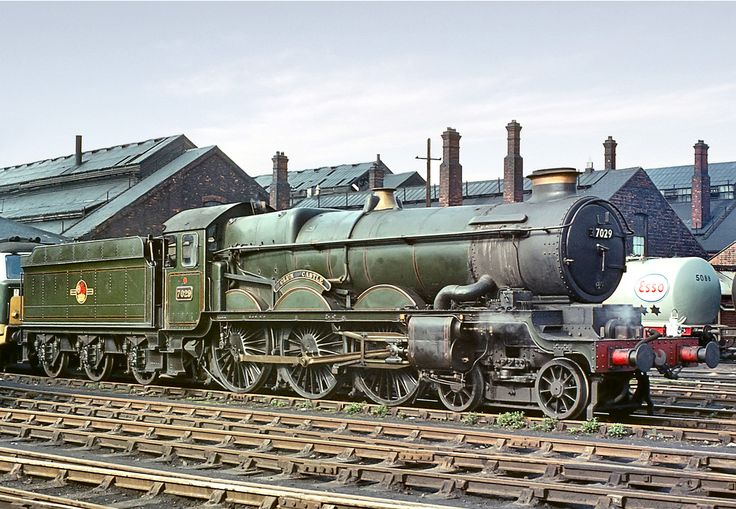 an old fashioned train is parked on the tracks in front of a building with chimneys