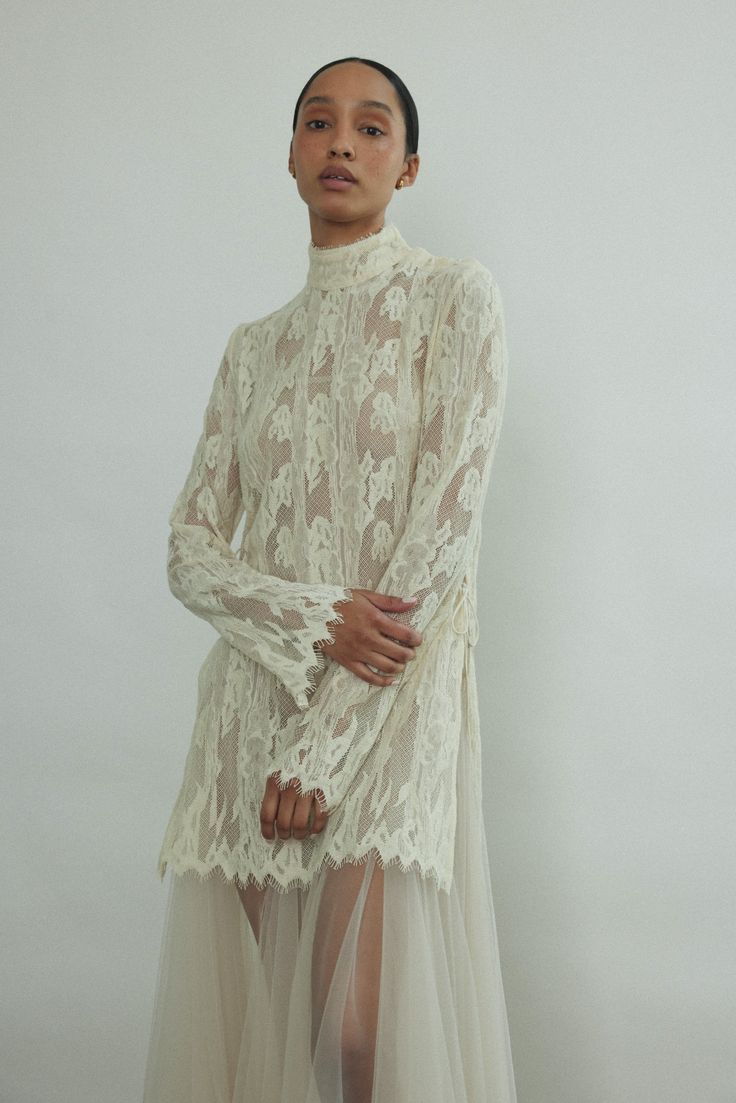 a woman standing in front of a white wall wearing a dress with sheer, lace detailing