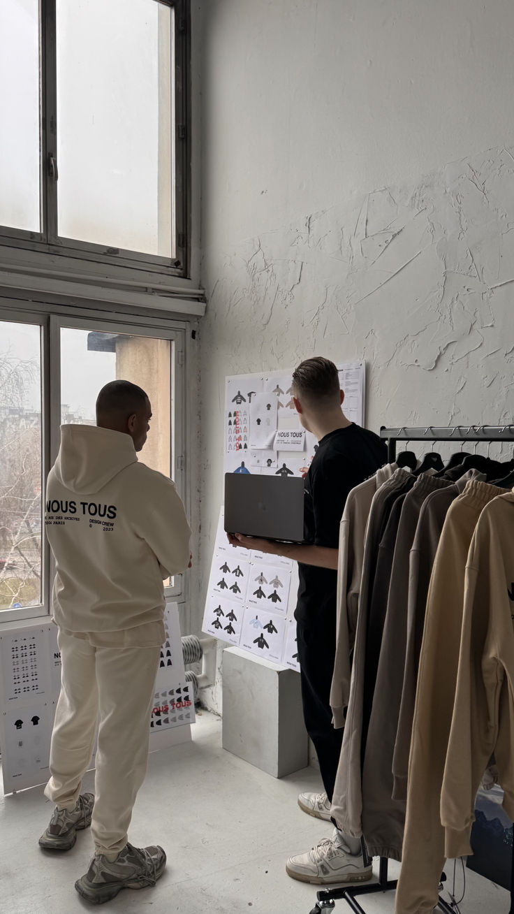 two men standing in front of a window looking at clothes on racks and hangers