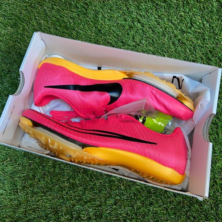 a pink and yellow nike shoe in a box on the grass with a tennis ball