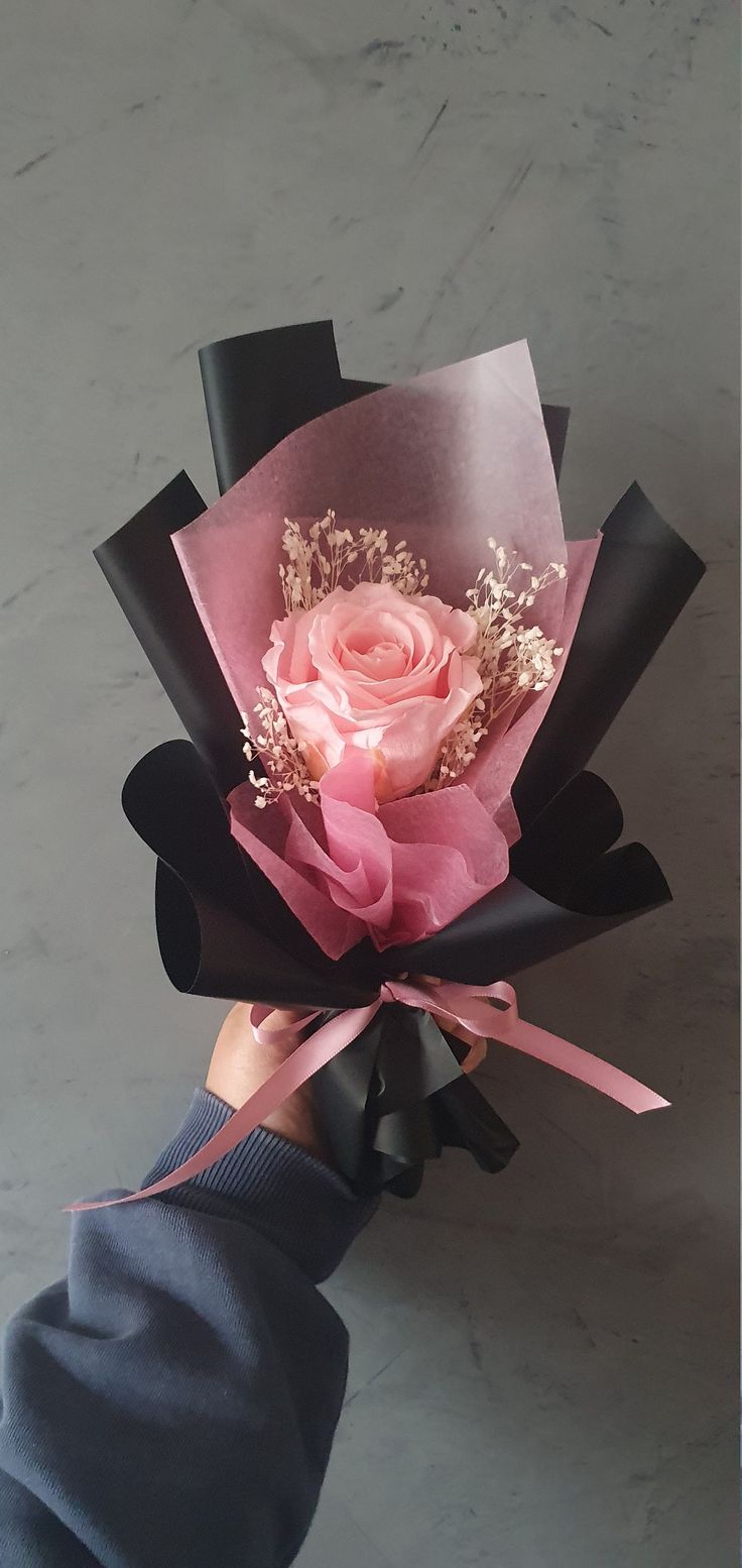 a hand holding a pink and black paper flower with ribbon around the end, on a marble surface