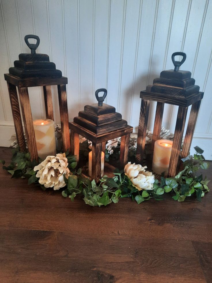 three wooden lanterns with flowers and greenery around them on a wood floor in front of a white wall