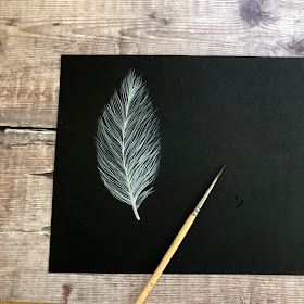 a piece of black paper with a white feather on it next to a paintbrush