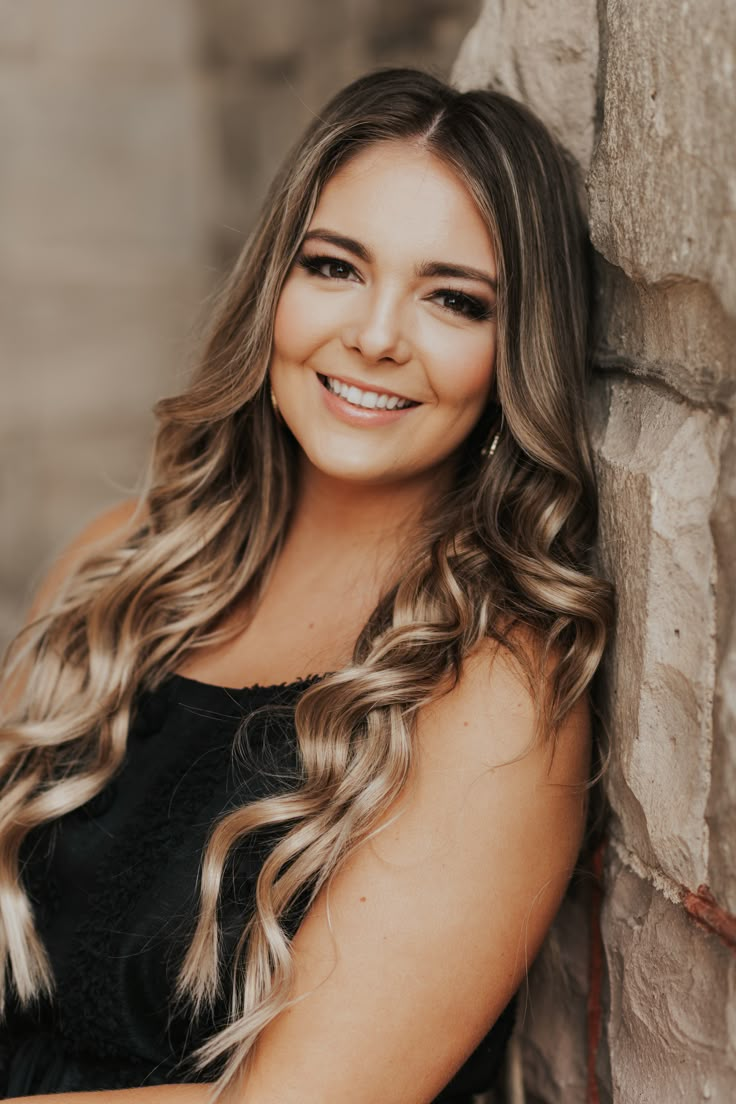 a woman with long hair leaning against a stone wall and smiling at the camera while wearing a black dress