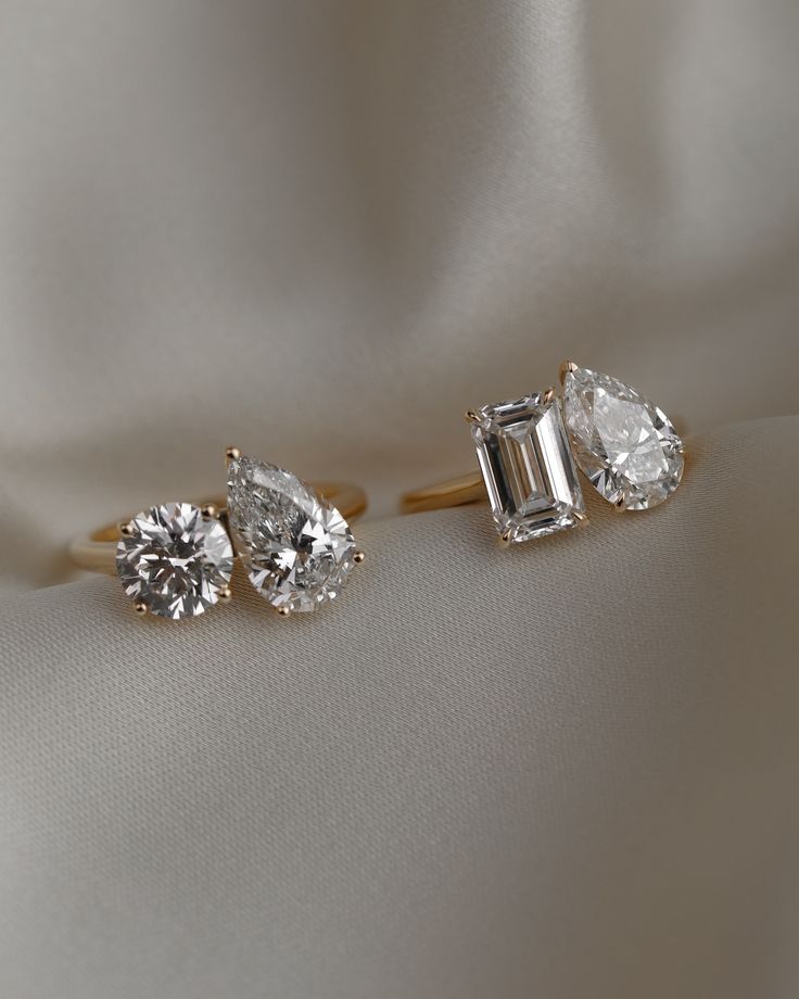 three diamond engagement rings sitting on top of a white satin material surface, with one ring in the shape of a baguette and two pear shaped diamonds