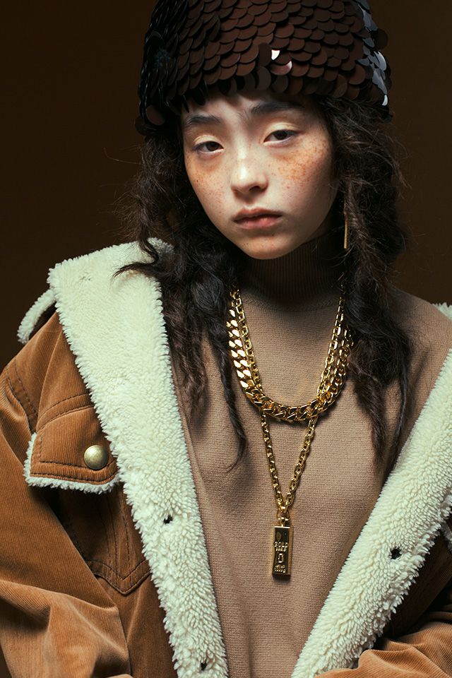 a woman with long hair wearing a brown coat and gold chains on her head, standing in front of a dark background