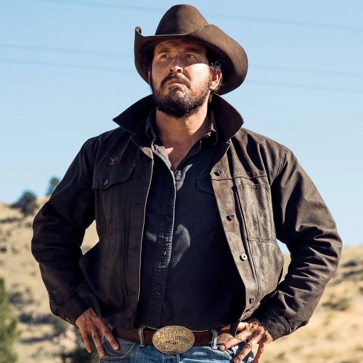 a man wearing a cowboy hat standing in front of a hill with his hands on his hips