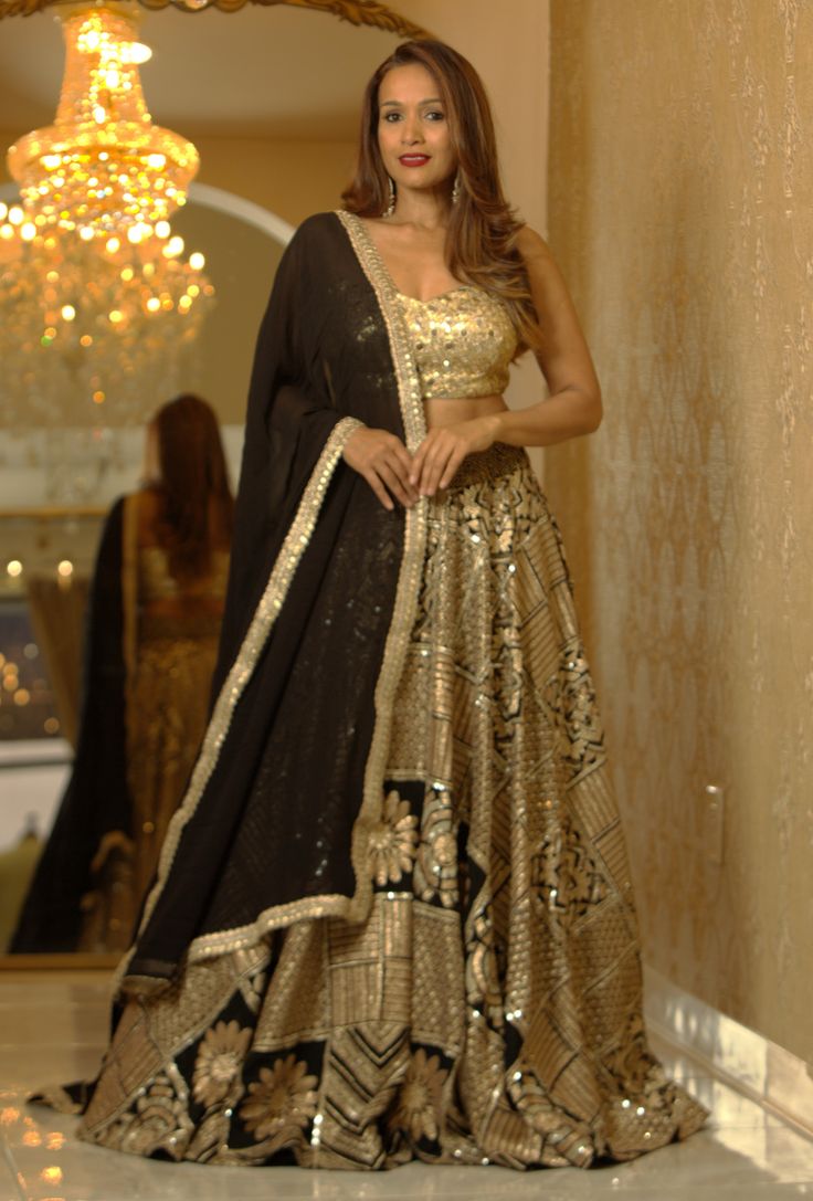 Experience the timeless allure of this enchanting Kanika Lehenga, adorned with intricate mirror, pearl, stone, and embroidery embellishments. Unleash your inner elegance and opulence with every step. Transform yourself into a vision of sophisticated royalty with each step in our timeless Kanika Lehenga. Adorned with a mesmerizing array of intricate mirror, pearl, stone, and embroidery embellishments, this luxurious piece is designed to unleash your inner elegance and opulence. Color - Black Gold Glamorous Tissue Silk Sets With Zari Work, Glamorous Tissue Silk Lehenga With Zari Work, Glamorous Tissue Silk Lehenga For Designer Wear, Hand Embellished Lehenga For Wedding And Transitional Seasons, Glamorous Chinon Lehenga For Festive Season, Glamorous Sets With Mirror Work In Chinon, Glamorous Chinon Sets With Mirror Work, Glamorous Gold Sets With Intricate Embroidery, Formal Lehenga With Sequins In Traditional Drape