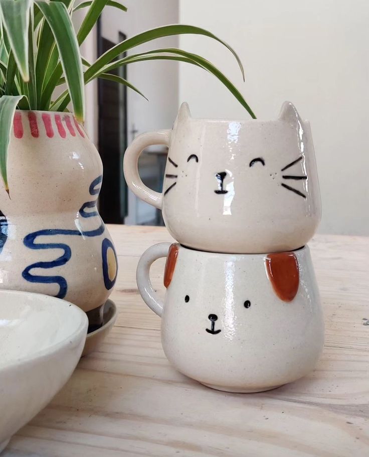 two ceramic cat mugs sitting next to each other on a wooden table with a plant in the background