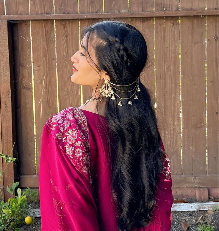 a woman with long black hair wearing a pink dress and gold jewelry stands in front of a wooden fence