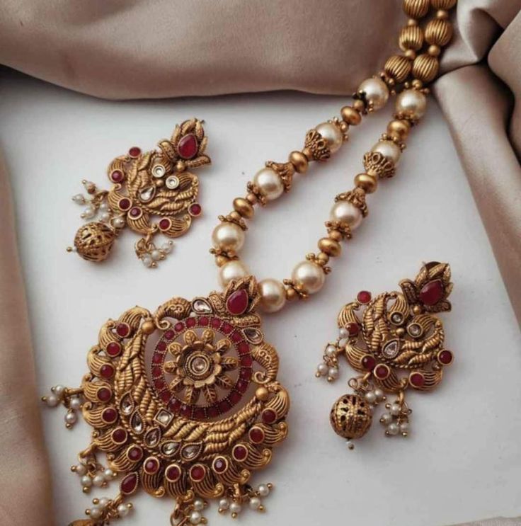 This beautiful Indian mala set features high-quality freshwater pearls and small, polished red gemstones, such as rubies or garnets, all elegantly set in a golden base. The necklace's traditional design showcases a symmetrical pattern of pearls interspersed with gemstones, enhanced by the golden setting and a central pendant. Matching earrings and a coordinated bracelet complete the set, echoing the necklace's luxurious style. Handcrafted by skilled artisans, the set combines premium materials a Elegant Kundan Jewelry Sets For Puja, Pearl Kundan Necklace With Intricate Design As A Gift, Festive Jewelry Sets With Pearl Pendant For Celebration, Traditional Pearl Bridal Necklace For Anniversary, Traditional Pearl Jewelry Sets, Elegant Hand Set Jewelry For Puja, Elegant Red Bridal Necklace For Puja, Elegant Round Kundan Necklace For Puja, Traditional Pearl Necklaces For Anniversary