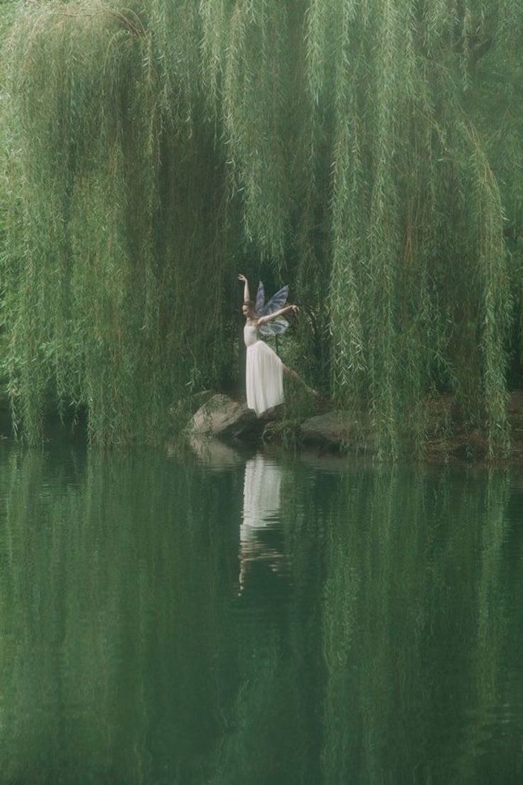 a woman in a white dress is standing on a rock by the water with willow branches