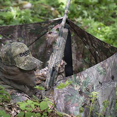 a man sitting in the woods with an umbrella over his head while holding a hunting gear