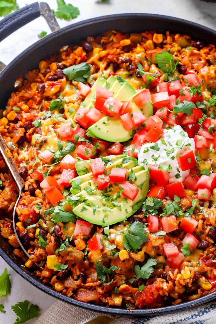 a skillet filled with mexican food and topped with avocado, tomatoes, cilantro, and sour cream