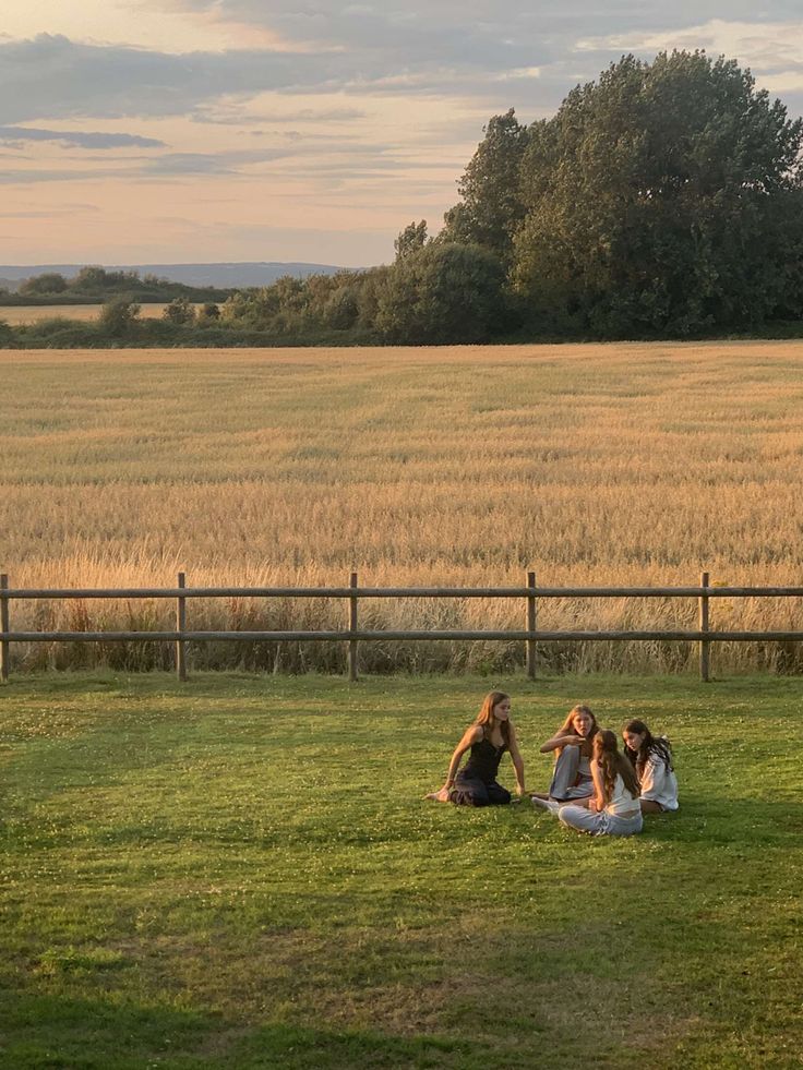 three women sitting on the grass in front of a large field with trees and fields behind them