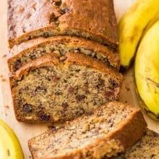 sliced banana bread sitting on top of a wooden cutting board next to two ripe bananas