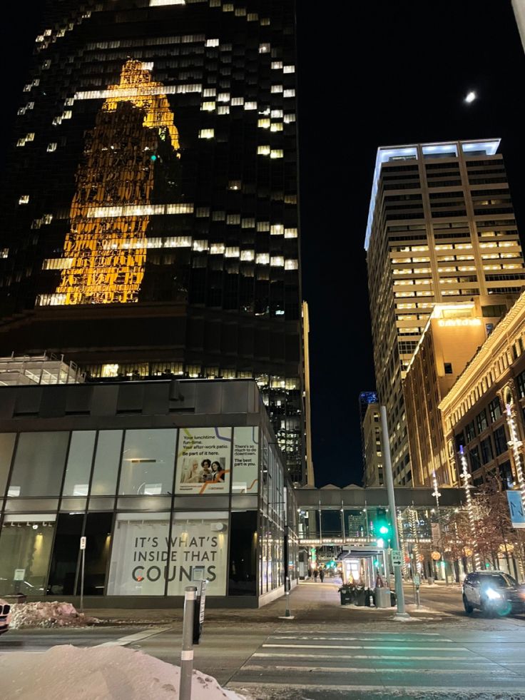 a city street at night with tall buildings lit up in the background and snow on the ground