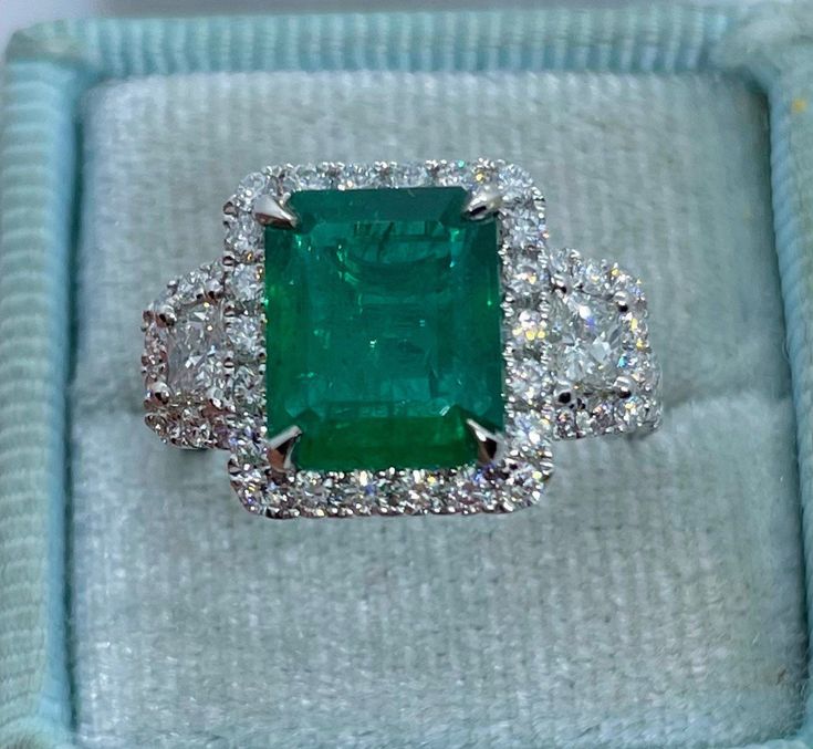 an emerald and diamond ring sits in a box on top of a cloth covered surface