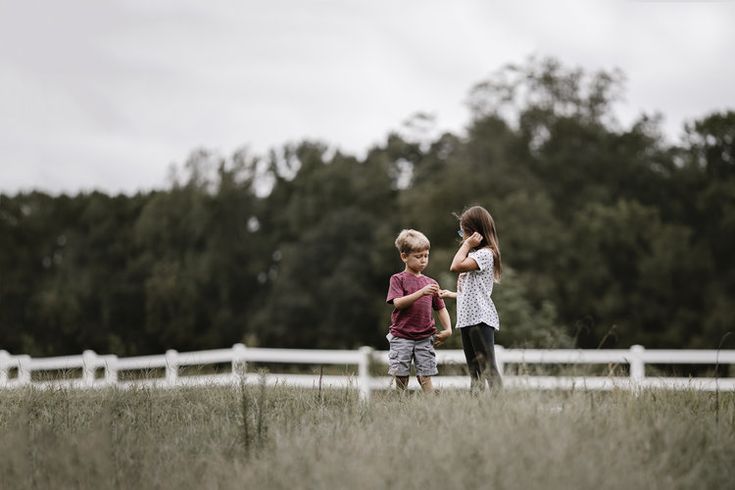 two young children standing in the grass near a white fence and looking at each other