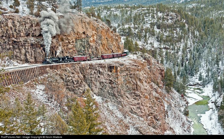 a train is coming down the tracks on a mountain side with trees and snow around it