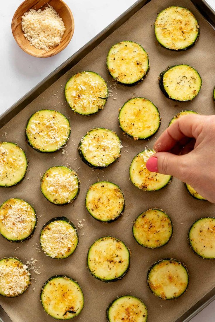 someone placing cheese on top of zucchini in a baking pan next to a wooden spoon