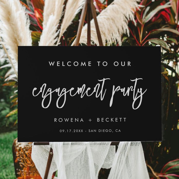 a welcome sign for an engagement party in front of some pamy plants and grass