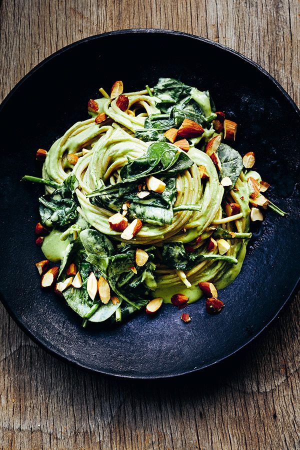 a black plate filled with pasta and greens on top of a wooden table next to a fork