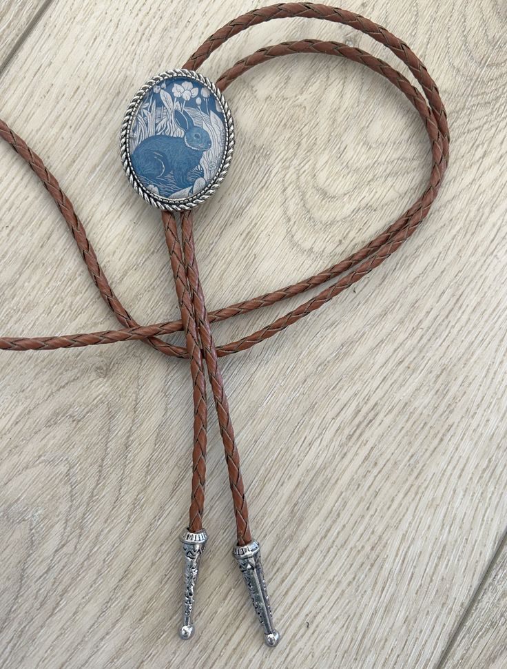 This awesome bolo tie has a vintage rabbit print with a clear resin coating and metal backing. Handmade in our shop!The cord is brown and made of genuine woven leather.Our bolos pair nicely with many of our belt buckles! They make wonderful gifts.The western bolo tie rope length is 39'' ; pendant size is 1 1/2'' x 1 1/4'' Handmade Lariat Bolo Tie For Rodeo, Vintage Lariat Bolo Ties For Rodeo, Handmade Lariat Bolo Tie For Western-themed Events, Vintage Bolo Ties With Adjustable Length For Rodeo, Vintage Bolo Ties With Adjustable Length For Western-themed Events, Vintage Bolo Tie With Adjustable Length For Western Events, Vintage Lariat Bolo Tie For Ranch, Vintage Lariat Bolo Ties For Ranch, Western Style Adjustable Bolo Ties