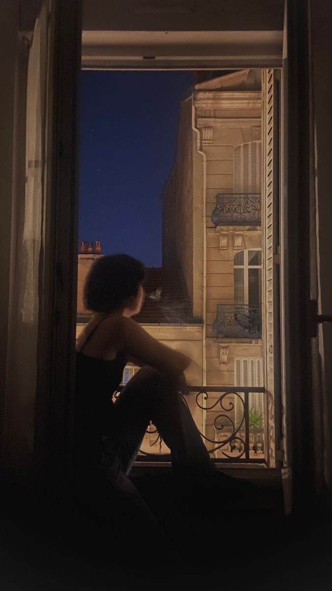 a woman sitting on a window sill looking out at the night sky and buildings