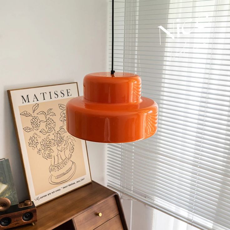 an orange lamp hanging from a ceiling in front of a window with blinds on it