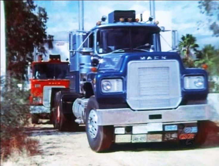 two large trucks parked next to each other on a dirt road with trees in the background