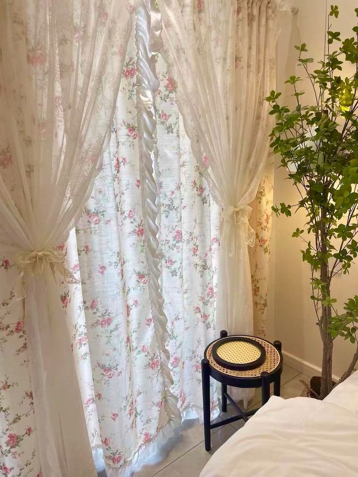 a bed sitting next to a window covered in white curtains and pink flowers on it