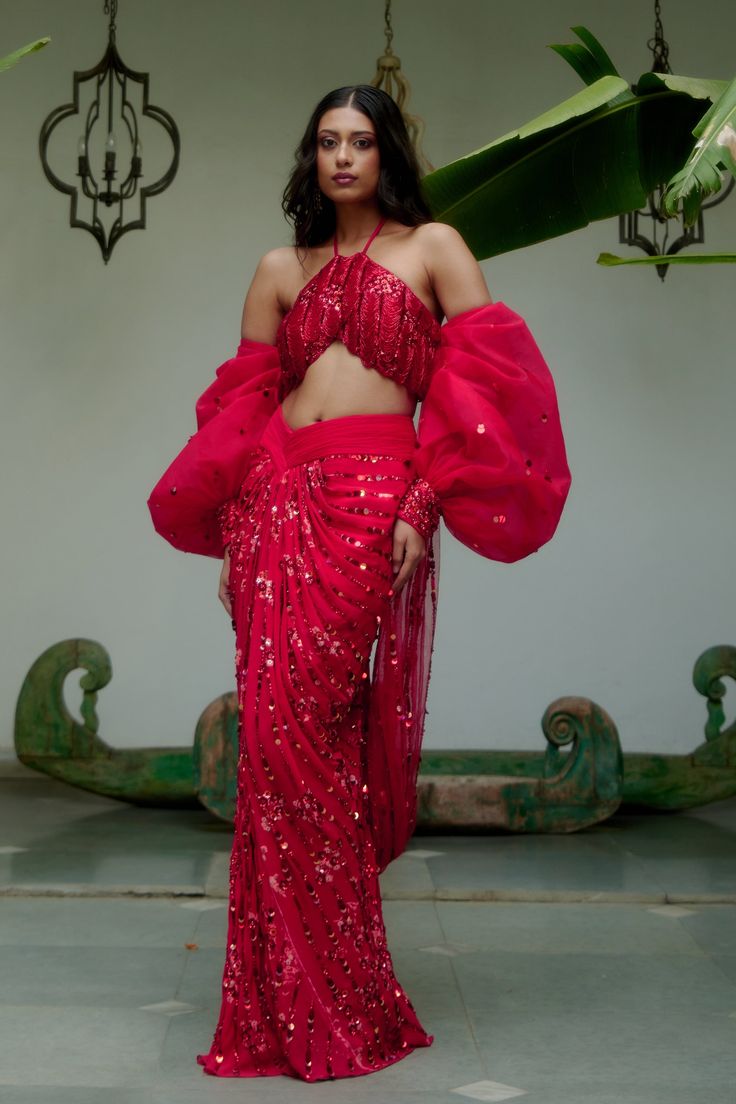 Step into iconic glamour where elegance is draped in the shimmer of scarlet palette for the season’s soirees. Our new-age concept of pre-draped saree features a chic halter neck blouse adorned with nalki crystals embellishment paired up with a sequined pre-draped skirt. Adding a hint of drama the handmade puffer sleeves complete the whole iconic look. • Pre-draped sequined skirt.• Halter-neck blouse adorned with nalki crystals and moti embellishments. • Metal hook at the back side of the blouse to conceal. • Puffed sleeves with an attached sequined dupatta.• Metal zip on the backside of the skirt along with a hook for support.From Moledro’s Fitoor collection. DELIVERY TIMEPlease allow 8-12 weeks for your outfit to arrive. FABRIC DETAILSBlouse: Butterfly Net Skirt: GeorgetteSleeves: Net Pro