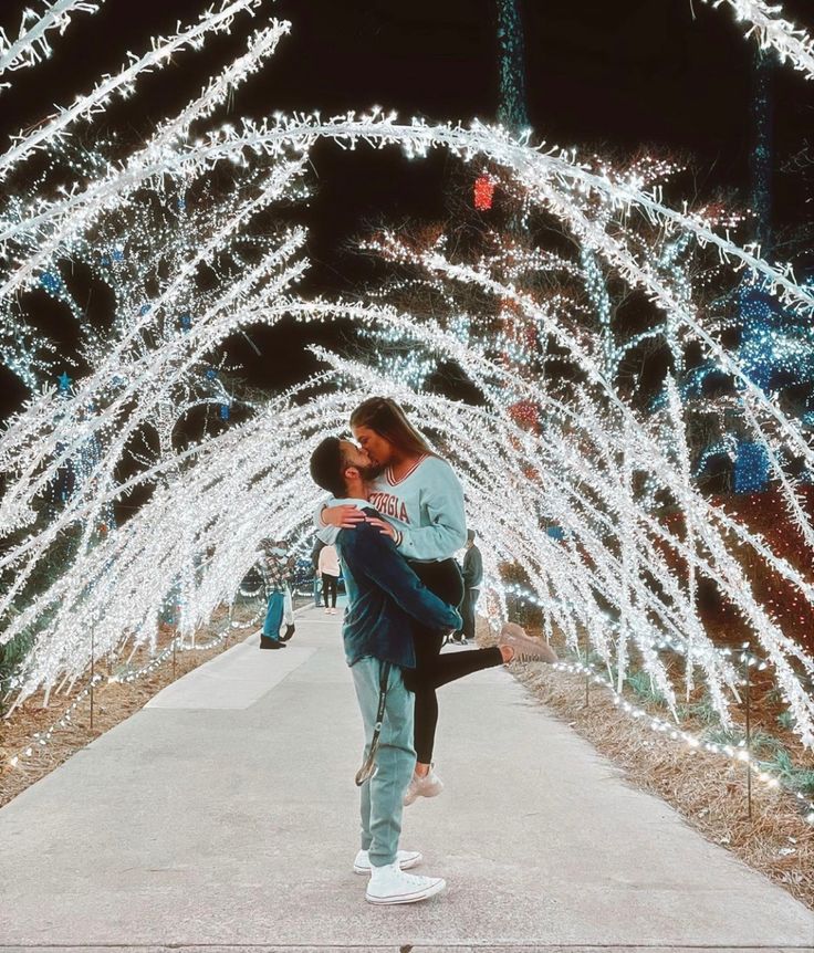 a man and woman kissing in front of christmas lights on the walkway at night time